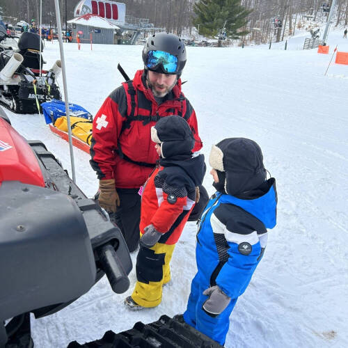 Ski patrol member Patrick Smith speaking to two children on a snowy ski slope near a snowmobile.